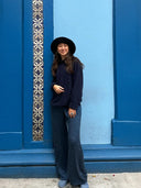 A woman in a black hat, dark sweater, and Rimmba | Sustainable Clothing Nindy Silk Pant - Indigo smiles as she stands against a bright blue wall with a decorative tile strip.