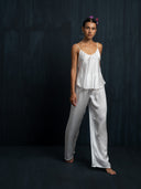 Barefoot woman wearing a white silk long pants and white camisole against a dark background.