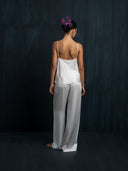 Barefoot woman wearing a white silk long pants and white silk camisole is facing a dark background.