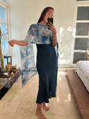 A woman stands barefoot in a sunlit bedroom, taking a mirror selfie while wearing a dark blue dress and the Rimmba | Sustainable Clothing Ocean & Waves - Indigo silk scarf, handcrafted from mulberry silk. A bed and table appear in the background.