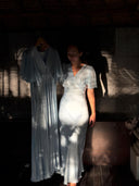 A woman in a flutter-sleeved Olivia Vintage Silk Dress - Aquamarine by Rimmba | Sustainable Clothing stands beside the same dress on a hanger, both partly lit by dappled sunlight and shadow for a dramatic effect.