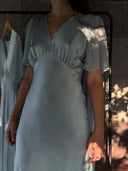 A woman wearing the Rimmba Olivia Vintage Silk Dress in Aquamarine, crafted from mulberry silk satin, stands indoors as sunlight and shadows highlight her. Behind her, a similar Rimmba sustainable dress hangs on a rack.