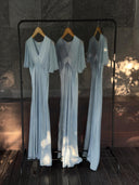 Three Olivia Vintage Silk Dresses in Aquamarine by Rimmba | Sustainable Clothing hang side by side on wooden hangers, their mulberry silk satin softly gleaming against a dark stone wall as sunlight and shadows create a dappled effect.
