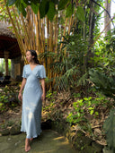 A woman wearing the Rimmba Olivia Vintage Silk Dress in Aquamarine stands barefoot on a stone path amid lush greenery and bamboo, gazing to her left.