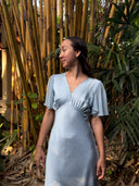 A woman in the Rimmba Olivia Vintage Silk Dress - Aquamarine stands before tall bamboo and lush greenery, gazing left with a calm expression. Sunlight streams through foliage, highlighting the dress’s sustainable elegance.
