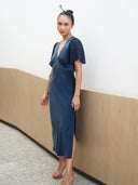 A woman in an indigo Olivia Vintage Silk Dress by Rimmba | Sustainable Clothing, made from mulberry silk satin with short sleeves and a V-neck, stands indoors by a white textured wall and wooden panel, looking at the camera in clear heeled sandals.