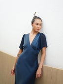 Woman with hair in a bun wears the Olivia Vintage Silk Dress - Indigo by Rimmba | Sustainable Clothing, featuring short flutter sleeves. The elegant mulberry silk satin shimmers as she stands against a white textured wall and light wood panel.