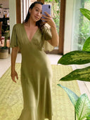 A woman wearing the Olivia Vintage Silk Dress - Mustard Green by Rimmba | Sustainable Clothing takes a mirror selfie in a bright, vintage-style room with large windows, lush plants, and a patterned rug.