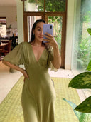 A woman takes a mirror selfie in a bright room with plants, wearing the Olivia Vintage Silk Dress - Mustard Green by Rimmba | Sustainable Clothing. Large windows and a green patterned rug add to the elegant, vintage-inspired scene.