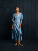 Barefoot woman wearing Mulberry silk midi dress baby indigo, v neckline with flowing silkchiffon sleeves. Handcrafted botanical-dyed midi dress sustainable luxury fashion for formal and resort wear.