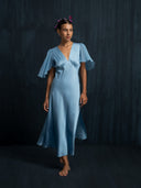 Barefoot woman wearing Mulberry silk midi dress baby indigo. Hancrafted botanical-dyed sustainable luxury fashion for formal and resort wear against a dark background.
