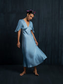 Barefoort woman wearing a flowing Mulberry silk midi dress baby indigo against a dark background. Handcrafted botanical-dyed sustainable luxury fashion for formal and resort wear.