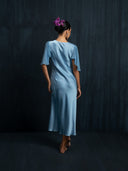 Back view of woman wearing Mulberry silk midi dress baby indigo with v-neck style and silkchiffon sleeves. Hancrafted botanical-dyed sustainable fashion for formal and resort wear.