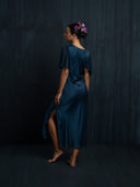 Back view of woman wearing Rimmba's Olivia vintage dress indigo. Hancrafted botanical-dyed sustainable luxury fashion for formal and resort wear. Hairbun style with purple flower against a dark background.