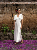 A woman in the Olivia Vintage Silk Dress - White by Rimmba | Sustainable Clothing stands barefoot on purple petals, against a rustic stone and earthen wall, gazing calmly to the side.