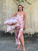 A woman in a sleeveless tie-dye maxi dress from Rimmba | Sustainable Clothing stands barefoot on pavement, holding the Pua Silk Pillow Case 2x - Set, posing before a weathered wall with greenery overhead.