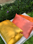 The Pua Silk Pillow Case 2x - Set from Rimmba | Sustainable Clothing, featuring one yellow and one orange hypoallergenic pillow, sits outdoors on a white surface near grass and leafy plants with purple flowers.