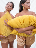 Two women wearing gold earrings and satin shorts stand side by side holding Rimmba’s Pua Silk Pillow Case 2x - Set against a white background. Both have long hair and neutral expressions; one looks ahead, the other gazes aside.