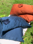 The Pua Silk Pillow Case 2x - Set by Rimmba | Sustainable Clothing features two hypoallergenic pillows, one rust orange and one dark blue with ruffled accents, displayed outdoors on a white surface atop green grass in sunlight.