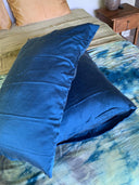 A large blue pillow with a hypoallergenic Pua Silk Pillow Case 2x - Set from Rimmba | Sustainable Clothing rests on yellow and blue tie-dye bedding, with tan pillows, a wooden nightstand, and a round decorative item in the background.