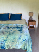 A neatly made bed with blue and green tie-dye bedding, dark blue Pua Silk Pillow Case 2x by Rimmba, and a beige sheet. A wooden nightstand with a textured lamp and book sits to the right against a light yellow wall.
