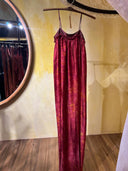 The Parvati Red-Eco Print Dress by Rimmba | Sustainable Clothing, a long, sleeveless mulberry silk dress with thin straps, hangs on a wooden hanger against a yellow wall with a round mirror and wooden floor in the background.