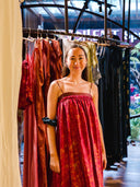 A woman in a red, sleeveless Parvati Red-Eco Print Dress by Rimmba | Sustainable Clothing smiles in a boutique, surrounded by colorful dresses on racks. Warm lighting and large windows fill the background with natural light.
