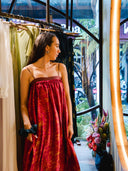 A woman in a Parvati Red-Eco Print Dress by Rimmba | Sustainable Clothing stands by a rack in a boutique adorned with botanical dyeing and fresh flowers, gazing out the window at motorcycles parked outside.