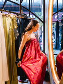 A woman stands in front of a mirror in a clothing store, holding out the sides of a bright red and gold patterned Parvati Red-Eco Print Dress by Rimmba | Sustainable Clothing, with other clothes hanging on nearby racks.