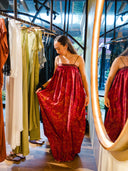 A woman stands in front of a mirror, holding the Parvati Red-Eco Print Dress by Rimmba | Sustainable Clothing, admiring its flowing Mulberry silk as she browses clothes on a rack in a stylish boutique.