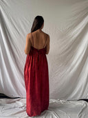 A woman with long dark hair stands facing away, wearing the Rimmba Parvati Red-Eco Print Dress, a backless, floor-length mulberry silk gown, in front of a white fabric backdrop.
