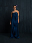 An indigo silk  maxi dress with thin straps and gathered bust detail. Front view against a dark background.