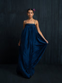 women wearing flowy slik maxy dress indigo with thin straps and gathered bust detail against a dark background.