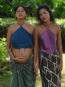Two women stand outdoors holding hands in Rimmba’s Pevita Halter Top – Mix Colors, one visibly pregnant. Their eco-conscious outfits reflect Rimmba’s sustainable values. Lush green foliage frames this serene moment.