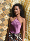 A woman with curly hair stands in sunlight against yellow and brown patterned fabric, wearing the Rimmba | Sustainable Clothing Pevita Halter Top - Mix Colors paired with a patterned skirt.