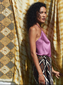 A woman with curly hair wears the Pevita Halter Top - Mix Colors by Rimmba | Sustainable Clothing, standing in sunlight before gold and brown patterned fabrics.
