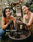 Two young woman smilling at the camera while working with a bucket of muddy water outdoors. Both of them have their hands covered in a dark mud.