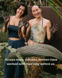 Two young woman smilling at the camera while working with a bucket of muddy water outdoors. Both of them have their hands covered in a dark mud.