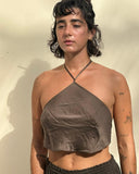 a young woman standing against a light-colored wall, illuminated by warm sunlight. She has short, dark, slightly wavy hair and is wearing large hoop earrings.She is dressed in a brown halter-style crop top made of a soft, slightly wrinkled fabric, paired with dark pants.