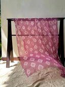 The Pink Shibori silk chiffon scarf by Rimmba | Sustainable Clothing, crafted with natural dyes, features a white geometric pattern and is draped over a dark wooden rack with soft sunlight casting gentle shadows on a beige backdrop.