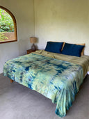 A neatly made bed features tie-dye green and blue bedding, three dark blue pillows with Rimmba's Pua Silk Pillow Case 2x - Set by Sustainable Clothing, a tan sheet, and sits near a window with a wooden frame and a nightstand with a lamp.