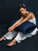 A woman was sitting holding a pillow covered in indigo silk wearing white cotton bandeau top and white cotton pants. hairbun style with purple flower against a dark background.