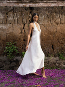A woman in a flowing white Aphrodite Dress by Rimmba | Sustainable Clothing walks barefoot on purple petals, set against a textured stone wall with green plants.