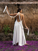 A woman wearing the Aphrodite Dress - White by Rimmba | Sustainable Clothing stands barefoot on purple petals, facing an old stone wall. She holds the dress’s white straps in both hands, her head turned slightly to the side.