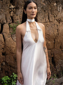 A woman with long dark hair and a floral tattoo on her upper arm stands by a rocky wall, wearing the Aphrodite Dress - White from Rimmba | Sustainable Clothing—a sleeveless, open-back silk dress with a choker.