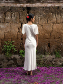 A woman in the Olivia Vintage Silk Dress - White by Rimmba | Sustainable Clothing, with a red flower in her hair, stands on purple petals before a textured brown stone wall lined with green plants.