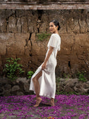 A woman wearing the Olivia Vintage Silk Dress - White by Rimmba | Sustainable Clothing stands barefoot on purple flower petals, posing sideways against a textured stone and brick wall with green plants.