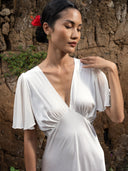A woman in the Olivia Vintage Silk Dress - White by Rimmba | Sustainable Clothing stands against a textured stone wall with eyes closed, gently touching her shoulder. She wears gold hoop earrings and has a red flower in her hair.