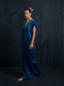 A woman stands barefoot against a dark background, wearing the Gaya Silk Kaftan Dress in indigo by Rimmba | Sustainable Clothing, featuring a V-neck and short sleeves. Her hair is styled back with pink flowers as an accessory.