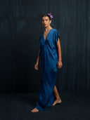 A woman stands barefoot against a dark background, gazing calmly to the side with a flower in her hair. She wears the Gaya Silk Kaftan Dress in Indigo by Rimmba | Sustainable Clothing.